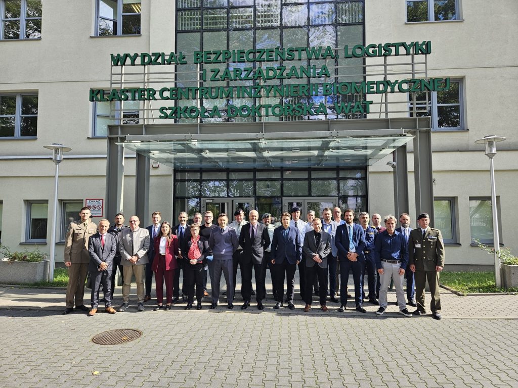Grupa kilkudziesięciu osób, w tym przedstawiciele w mundurach wojskowych oraz osoby w strojach formalnych, stoi przed wejściem do budynku Wydziału Bezpieczeństwa, Logistyki i Zarządzania, Wojskowej Akademii Technicznej. Nad wejściem widoczny jest napis: Wydział Bezpieczeństwa, Logistyki i Zarządzania, Klaster Centrum Inżynierii Biomedycznej, Szkoła Doktorska WAT.
